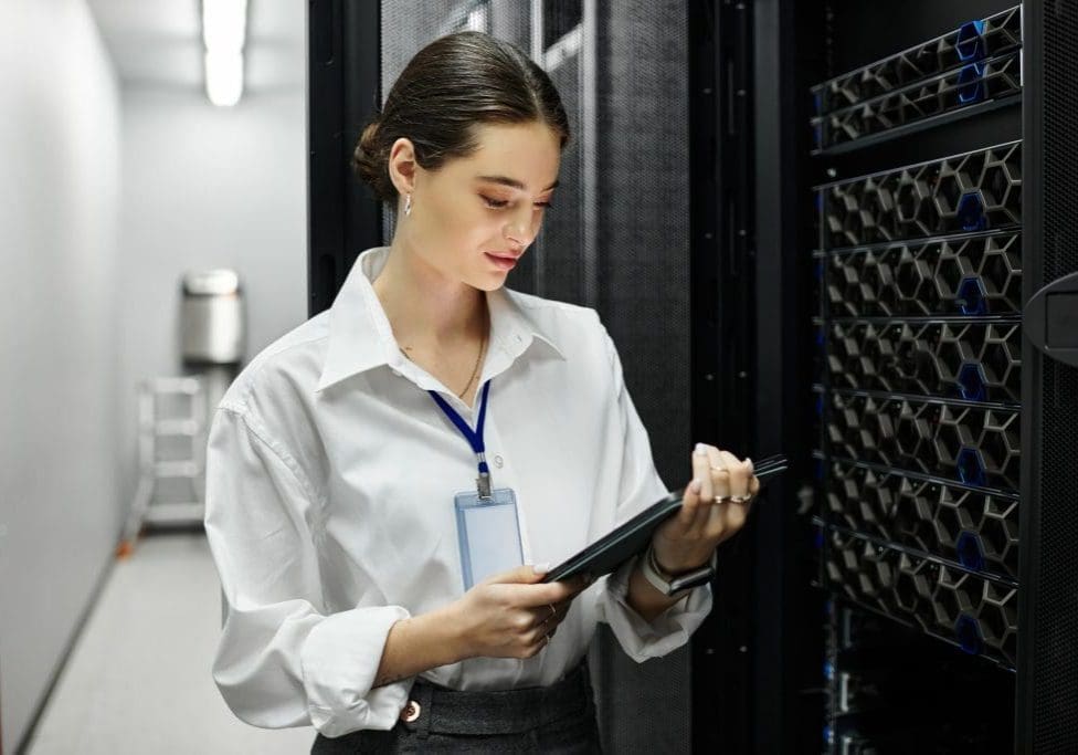 A woman in a white shirt is focused on her tablet while surrounded by server racks.