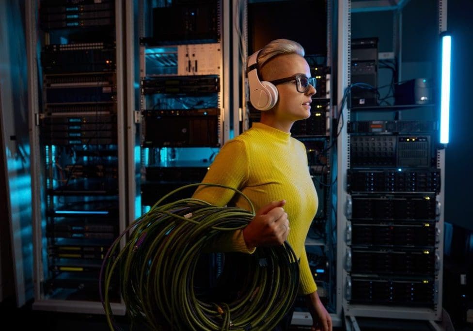 Young woman IT technician wearing headphones carrying wires bundle walking through server rack in big data center inside