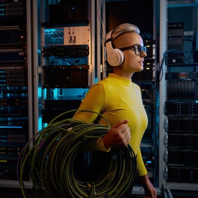 Young woman IT technician wearing headphones carrying wires bundle walking through server rack in big data center inside