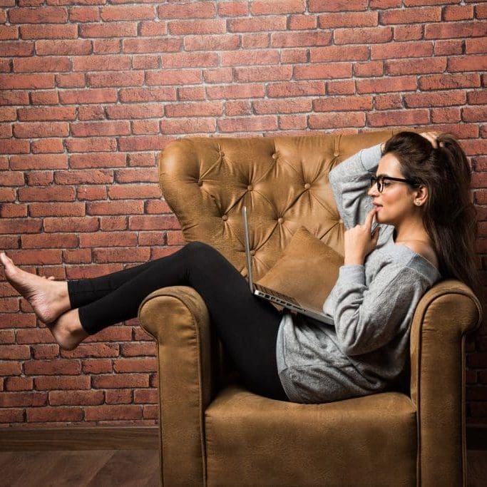 Indian girl with laptop sitting on wing chair against red brick wall