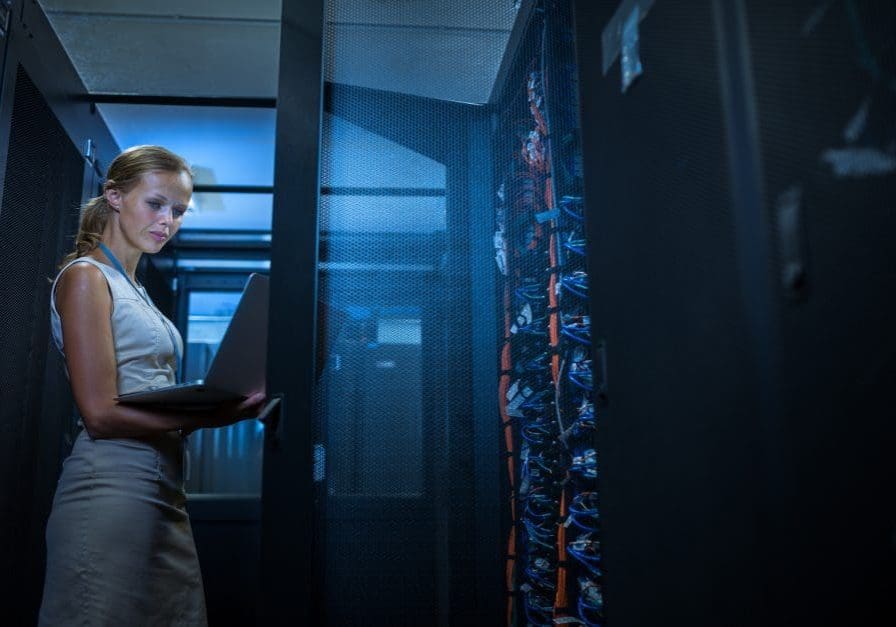 UK Servers IT engineer standing amid working server racks doing routine maintenance check and diagnostics using laptop computer