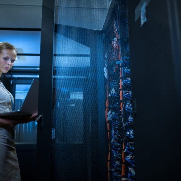 UK Servers IT engineer standing amid working server racks doing routine maintenance check and diagnostics using laptop computer
